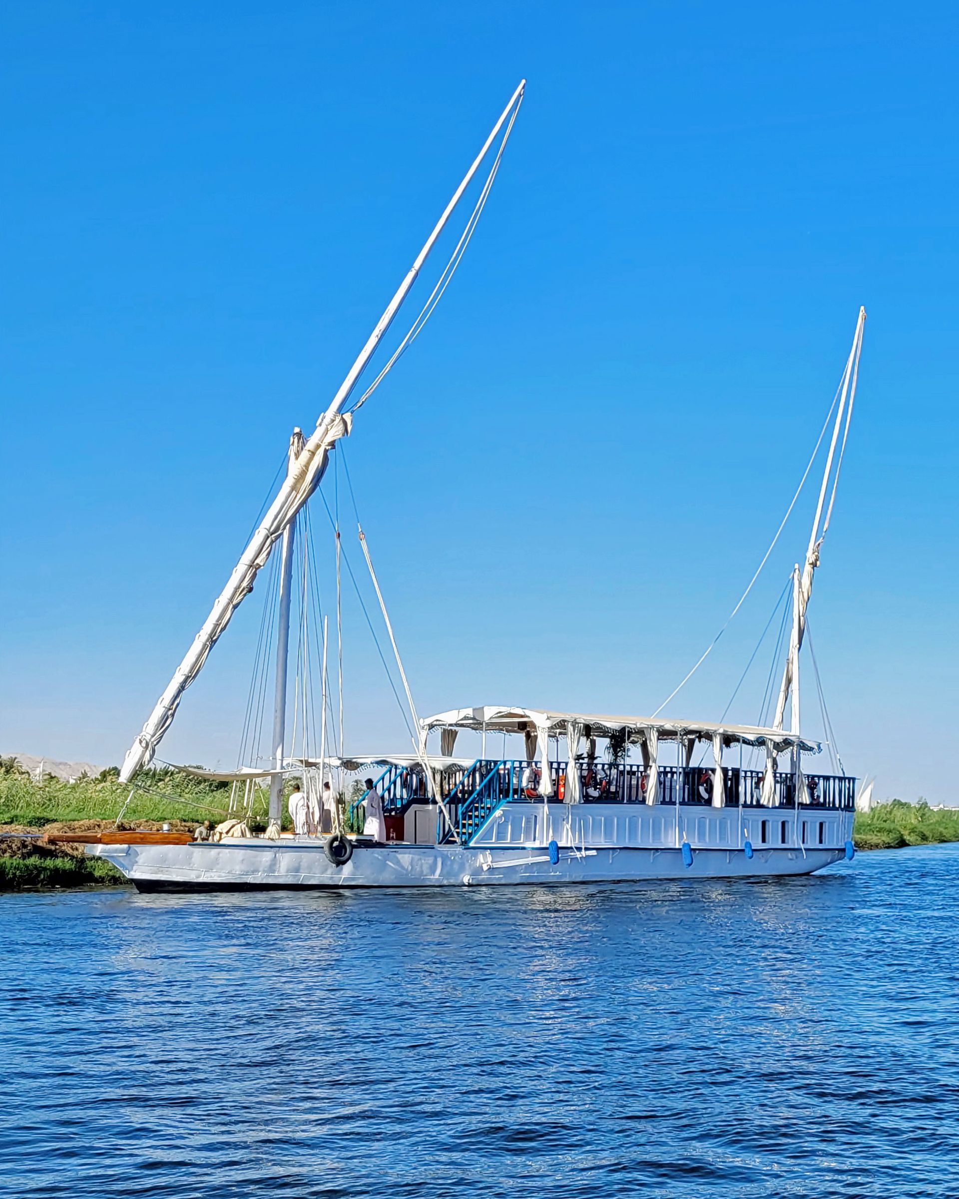 In this photo, you can see the Dahabiya Kingfisher, a traditional wooden boat that dates back to 1899. The boat has a graceful white hull, it sails along the Nile river, the longest and most ancient river in the world. The boat offers private cruises for travelers who want to experience the beauty and culture of the Nile valley, where they can visit temples, tombs, and villages along the way.