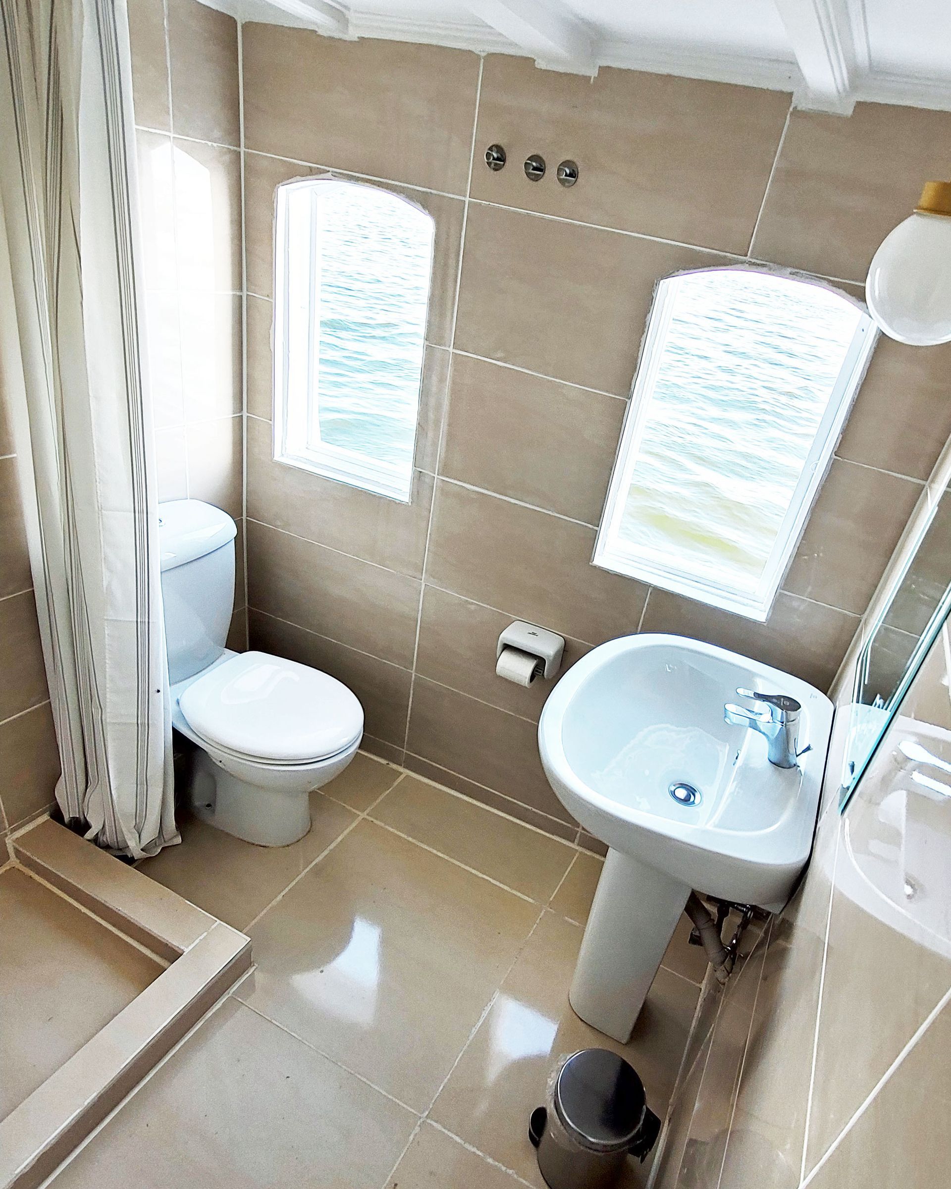 - A photo of a bathroom on a traditional Egyptian sailing boat
- The boat is called Dahabiya Kingfisher and dates back to 1899
- The bathroom has a white sink, a mirror, a shower and a window
- The window offers a view of the Nile river and the landscape