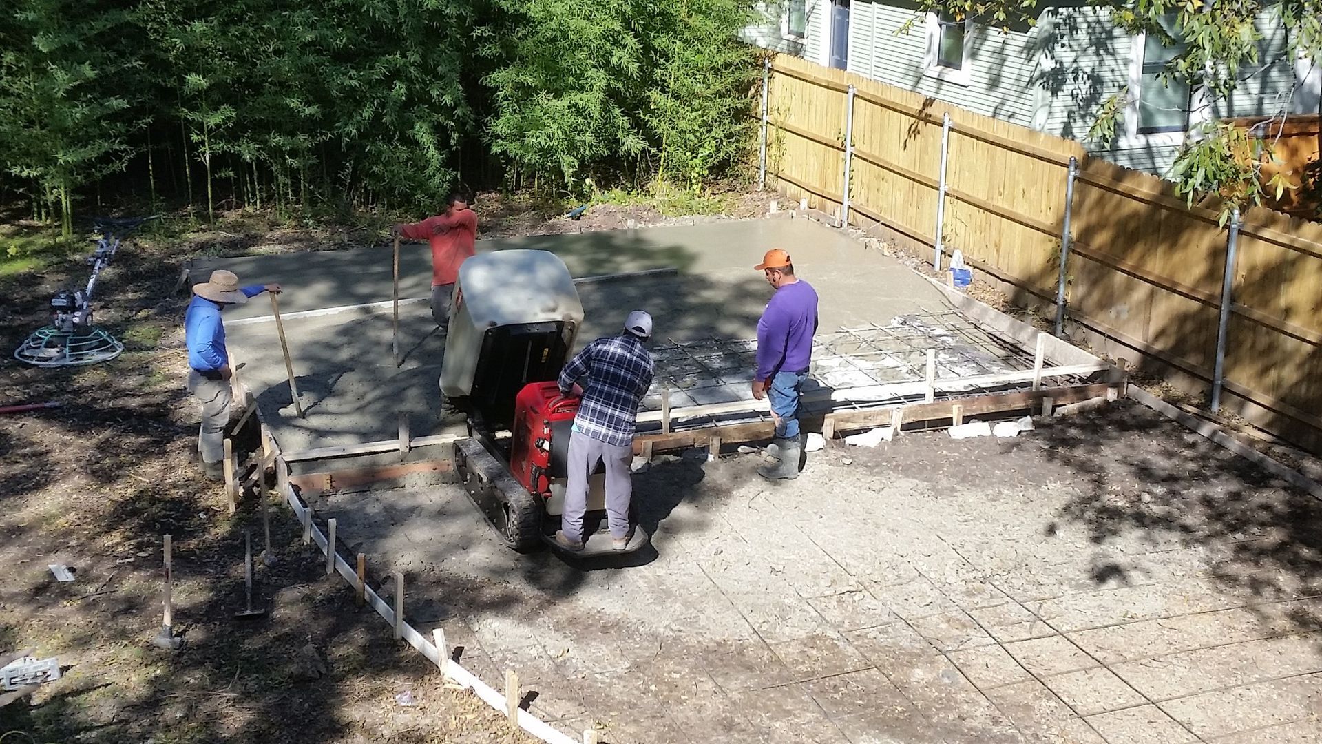 Construction workers pouring concrete, bordered by wooden forms and a fence, in a yard.