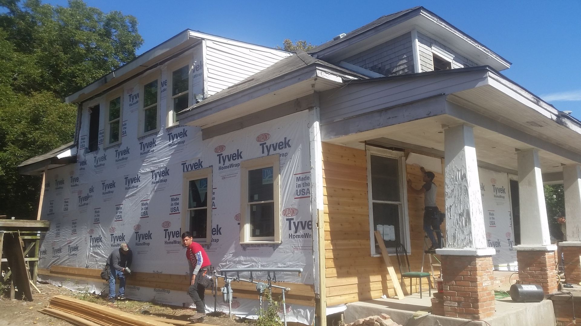 Construction workers siding a two-story house with white wrap and a covered porch on a sunny day.