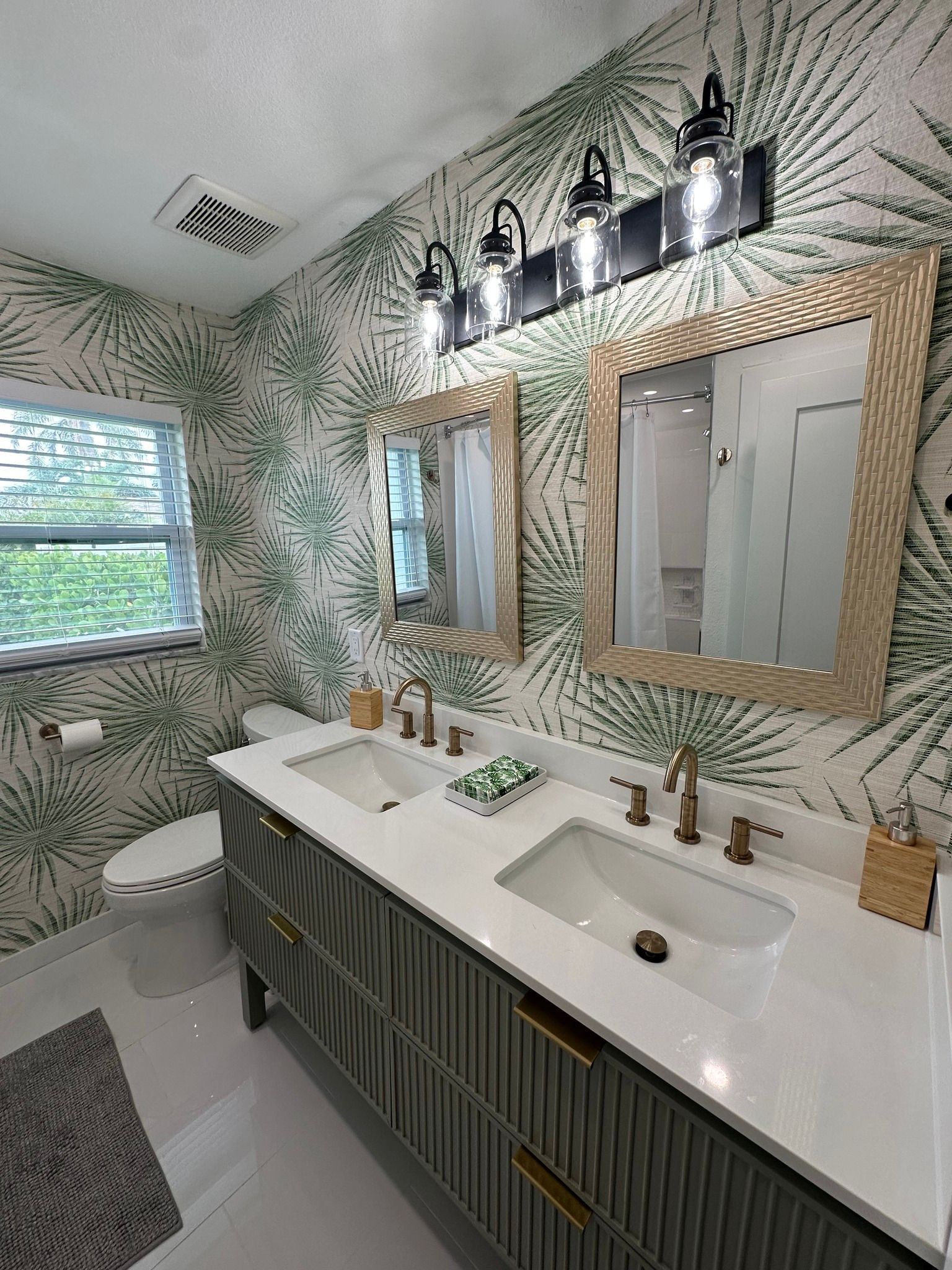 Bathroom with green leaf wallpaper, double sink vanity, gold-framed mirrors, and black light fixture.