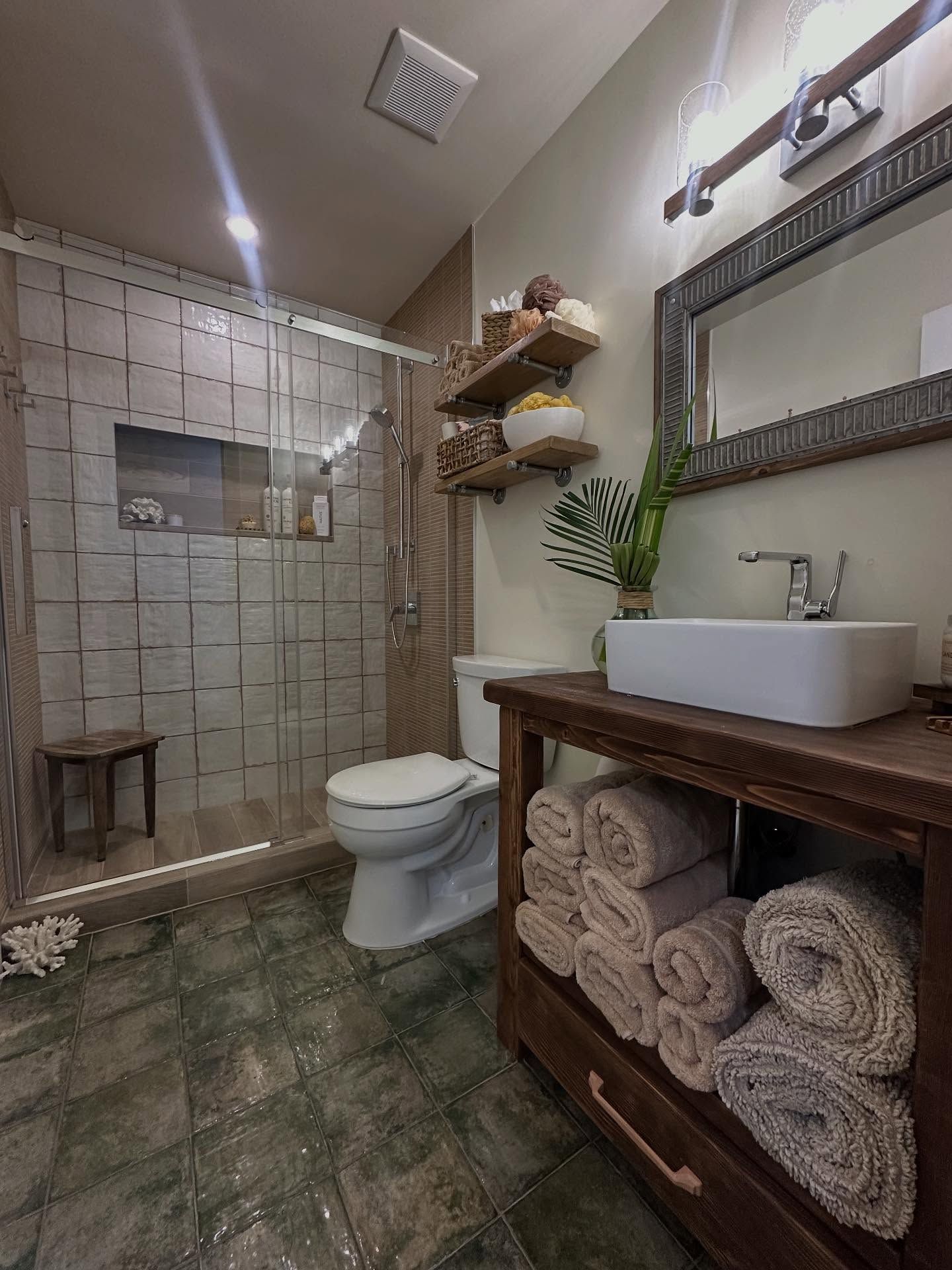 Rustic bathroom: glass shower, tiled walls, wooden vanity with towels, floating shelves.
