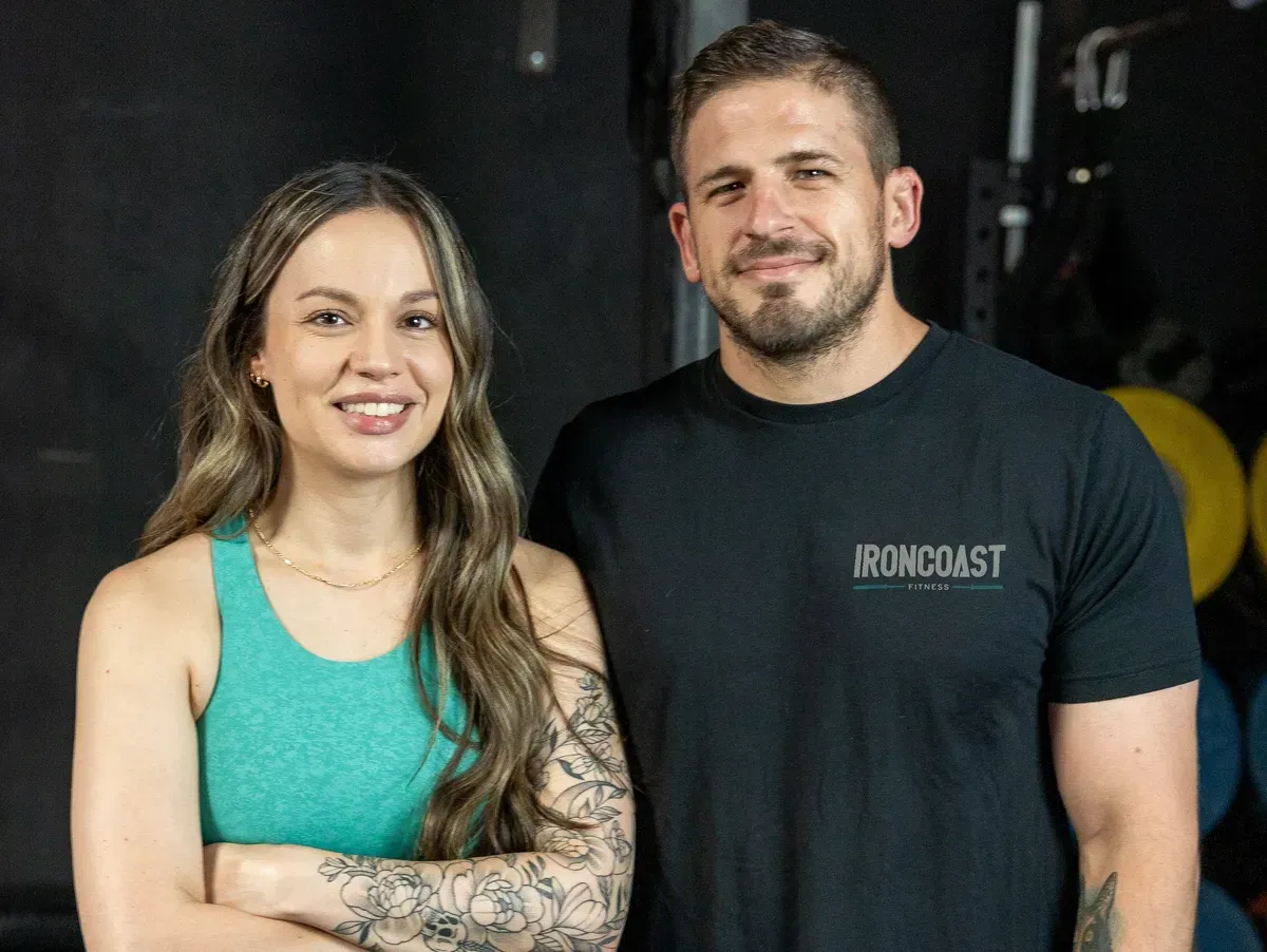 Woman and man smiling, in gym setting. Woman has arm tattoos, green top. Man in black shirt, arms crossed.