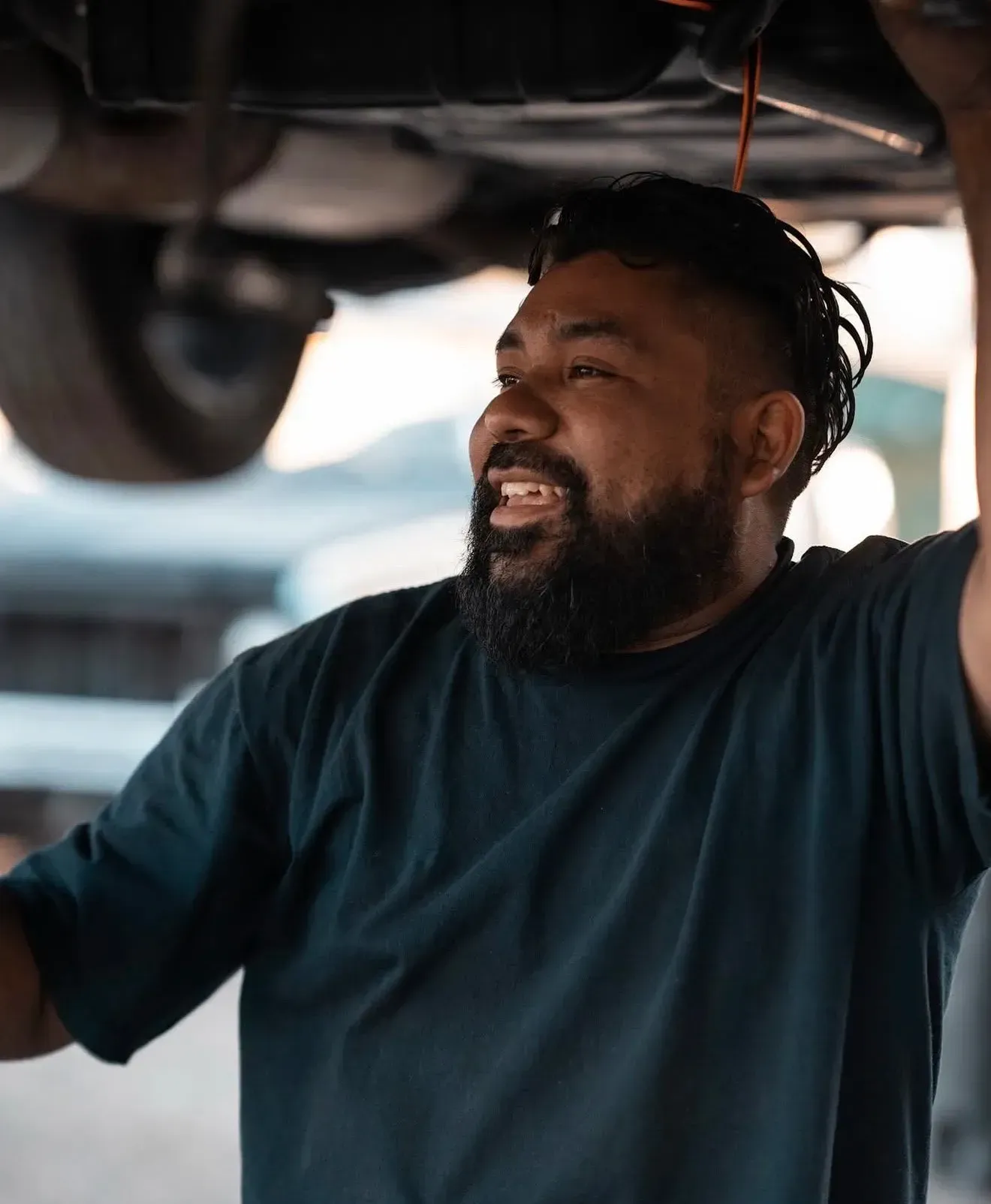 Mechanic smiling while working under a car, wearing a blue shirt | Nonstop Auto Reno, NV