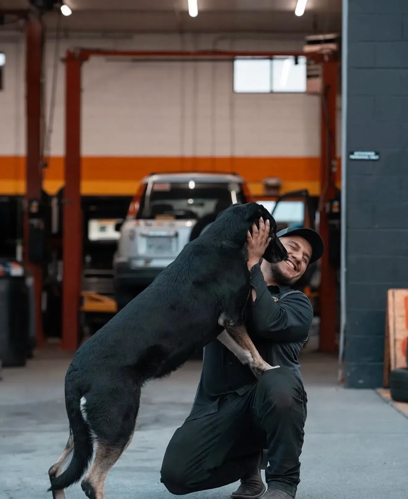 Mechanic kneels, embracing large black dog in a garage, smiling. Background: car, lift, and tools | Nonstop Auto Reno, NV