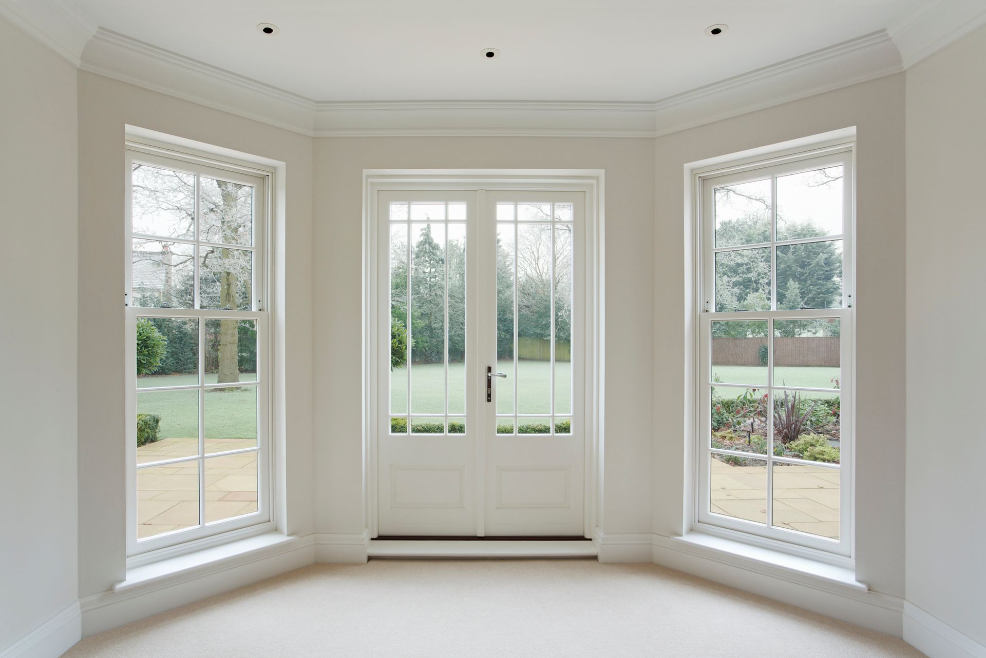 A bright, empty room with white walls, three windows, and double doors opening to a garden.