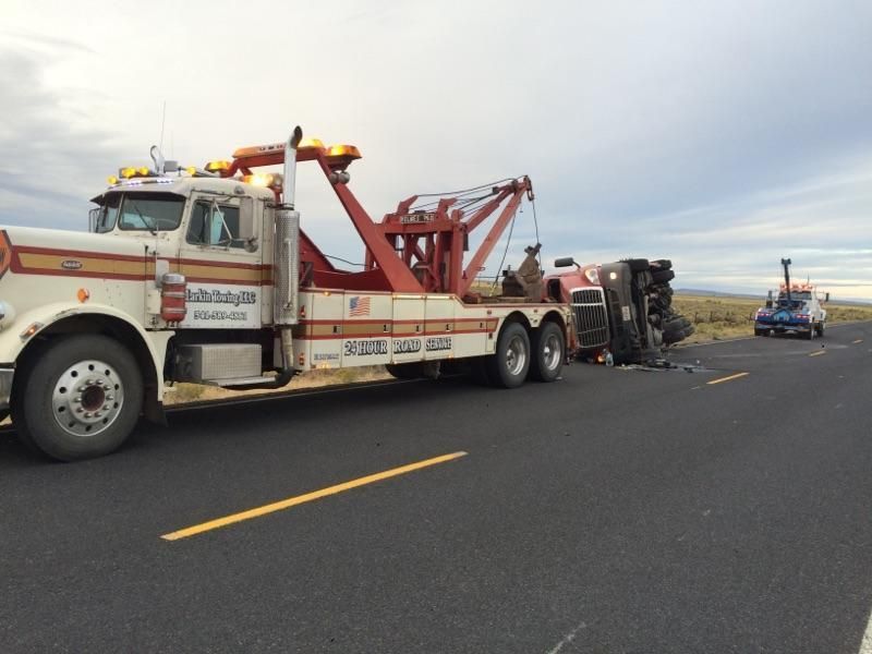 A large white and red tow truck sits on a rural road, connected to an overturned tractor-trailer in the distance.