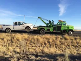 A green tow truck towing a white pickup truck along a rural roadside under a clear blue sky.
