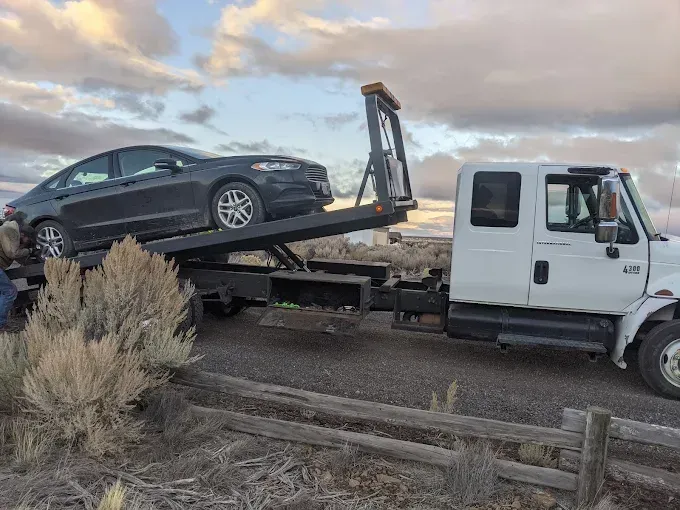 A dark sedan sits on the tilted flatbed of a white tow truck parked on a gravel roadside under a cloudy sky.