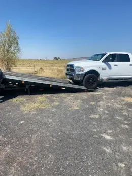 A white pickup truck is parked at the bottom of a lowered car hauler trailer on a gravel surface under a clear blue sky.