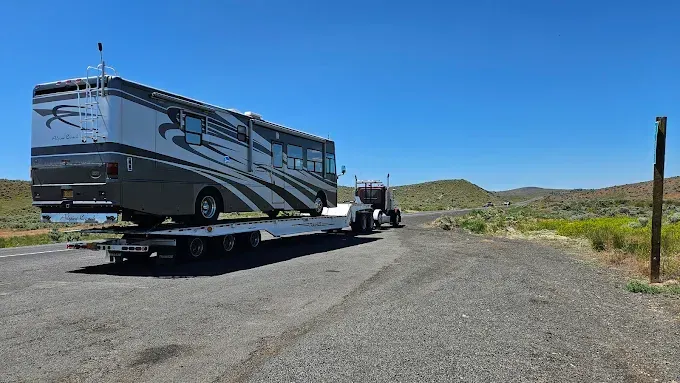 A semi-truck hauls a large RV on a flatbed trailer along a desert roadside under a clear blue sky.