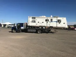 A large white travel trailer being transported on the flatbed of a blue truck in a gravel lot under a clear blue sky.