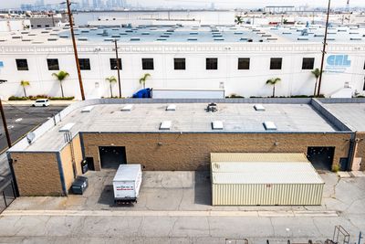 Warehouse exterior, brown brick building with a loading dock, white truck, and large white building in the background.