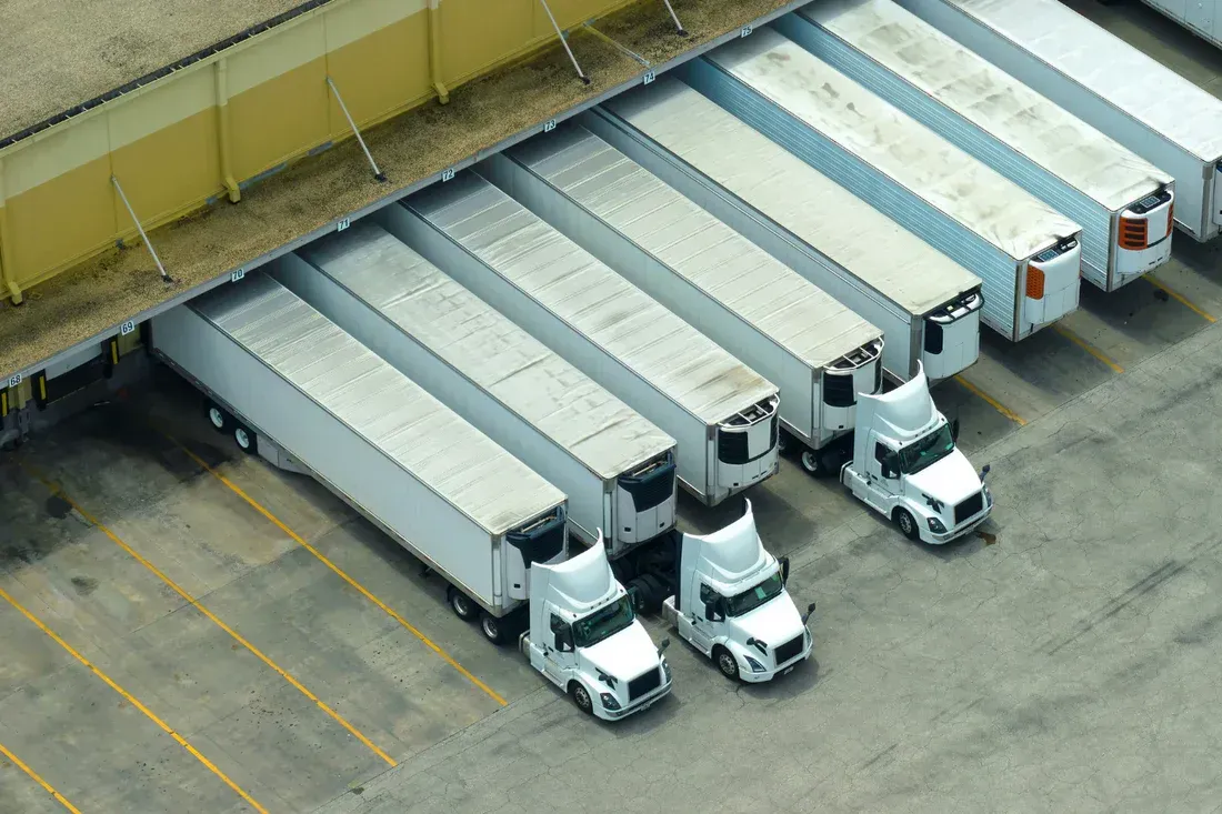 Trucks lined up at a loading dock, ready for loading/unloading. White trailers, yellow building, and concrete ground.