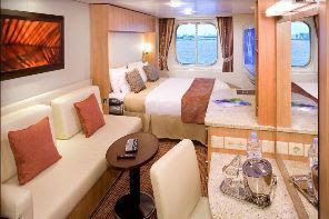 Cruise ship cabin, with bed, sofa, and small dining area. Natural light from porthole windows.