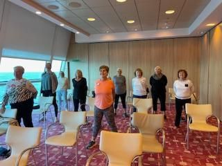 A group of people doing exercises in a well-lit room on a cruise ship, looking out at the ocean.