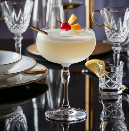 Cocktail in a fancy glass, garnished with cherry and orange, on a mirrored surface.