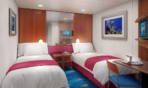 Cruise ship cabin with two twin beds, blue and red accents, desk, and artwork.