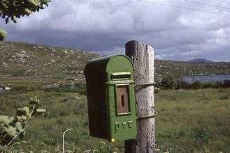 Green post box on a wooden post with a rural landscape background.
