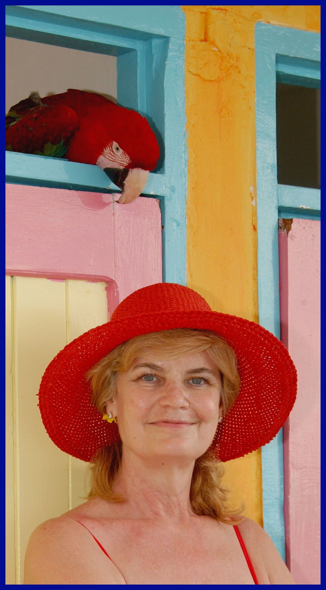 Woman in red hat smiles, red parrot peeks from colorful building.
