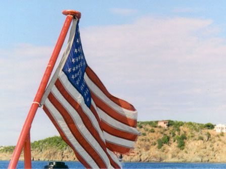 American flag waving on a red pole near the coast, against a blue sky and hillside.