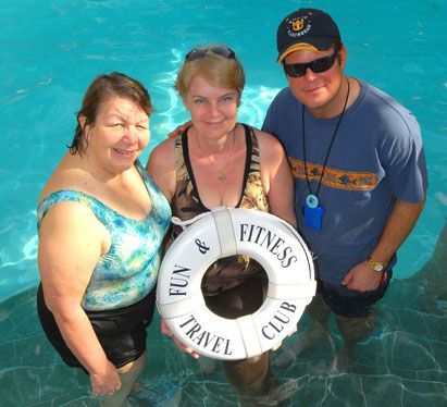 Three people in a pool, smiling. Woman in floral suit holds a life ring with 