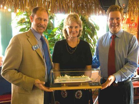 Three people holding a model boat in a restaurant. Woman smiles, men flank her, one with a blue tie.