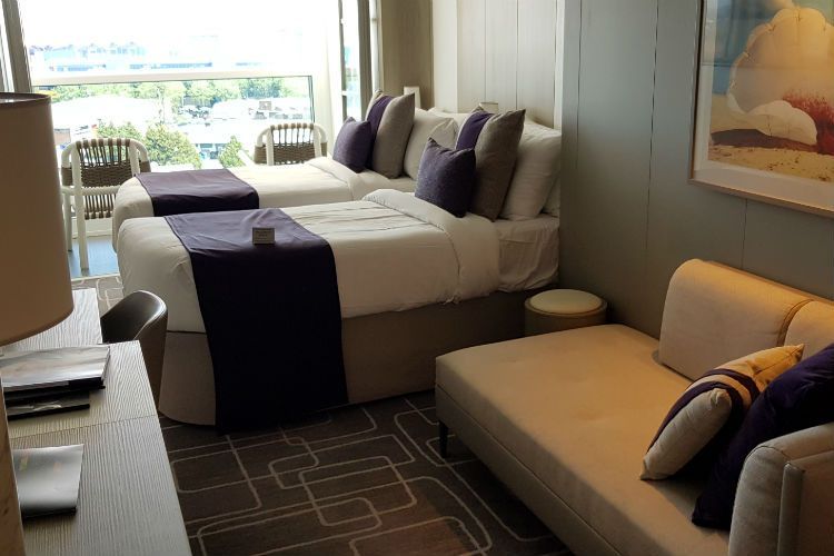 Cruise ship cabin with two twin beds, a sofa, and a balcony overlooking water.