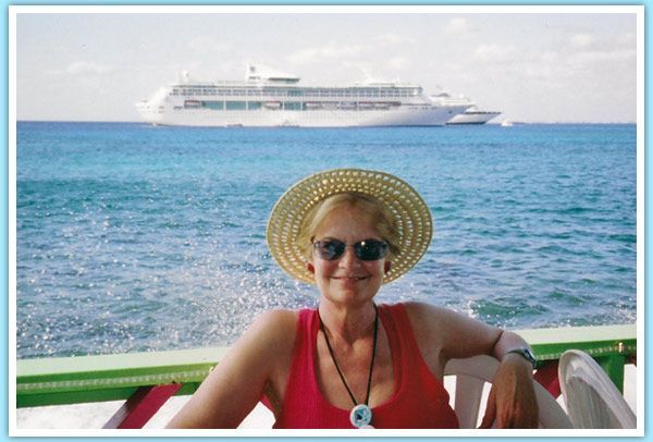 Woman in hat and sunglasses smiles, seated in front of a large cruise ship on a sunny day.