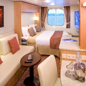 Cruise ship cabin interior: bed, small sofa, desk with ocean view, and a TV.