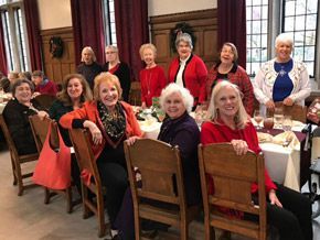 Group of women smiling at a table, some wearing red, indoors. Festive gathering with food and decor.