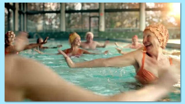 A group of seniors in a pool doing water aerobics. They're wearing swim caps, smiling and moving their arms.