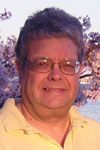 Man with glasses smiles. Pale yellow shirt, blurred background of trees.
