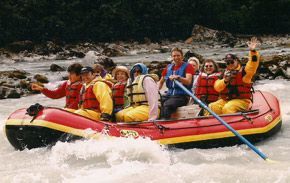 People rafting down a whitewater river in a red and yellow raft, waving.