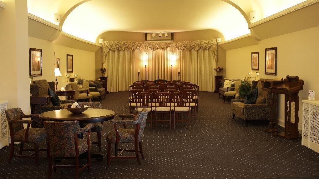 A formal, carpeted event hall with rows of chairs facing a curtained stage, flanked by small tables and seating areas.