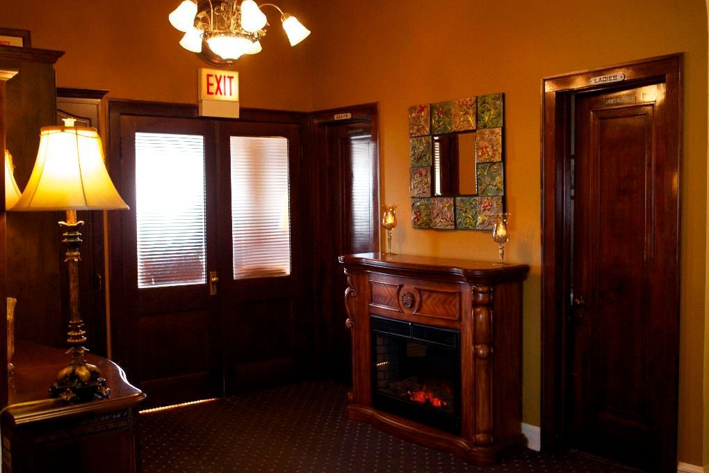 A dimly lit lobby featuring a fireplace, a lamp, an exit sign above wooden double doors, and a decorative mirror.