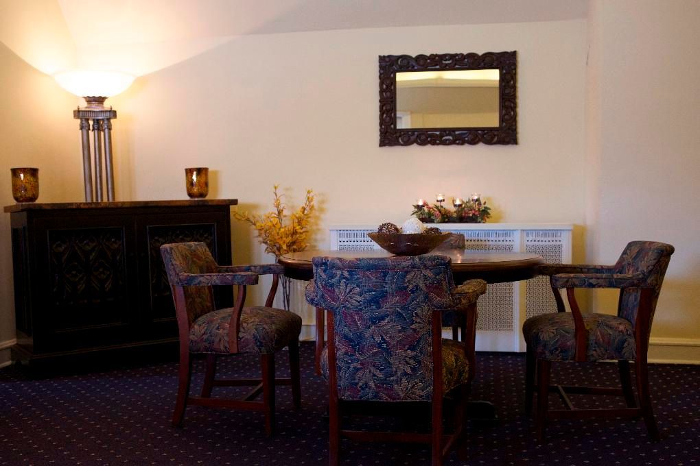 A dining area with a round wooden table, three patterned armchairs, a dark sideboard with a lamp, and a wall mirror.