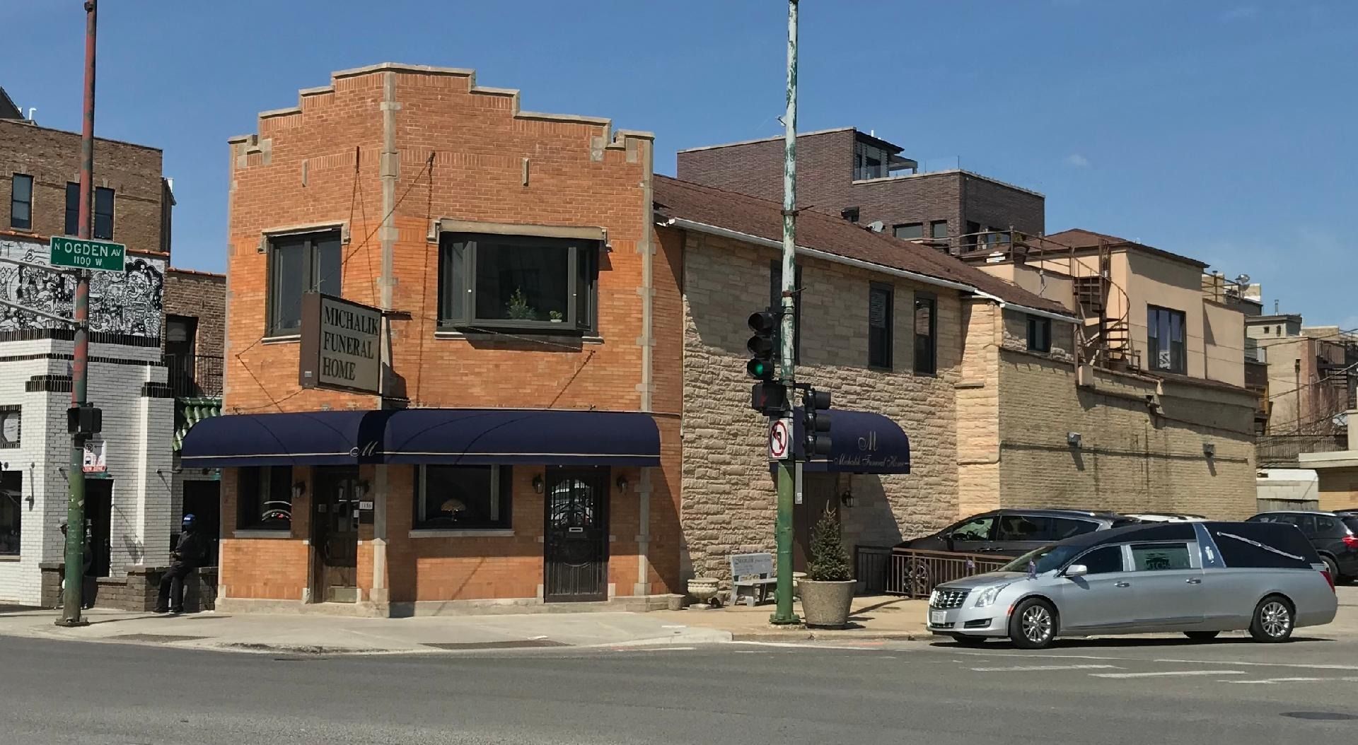 Brick building with blue awnings; hearse parked in front.