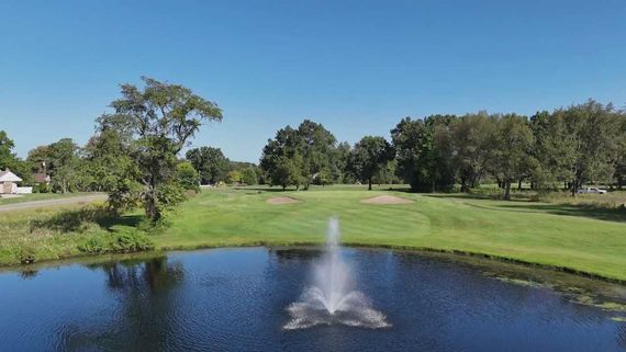 A golf course fairway featuring a central pond with a spraying fountain, bordered by green grass and mature trees.