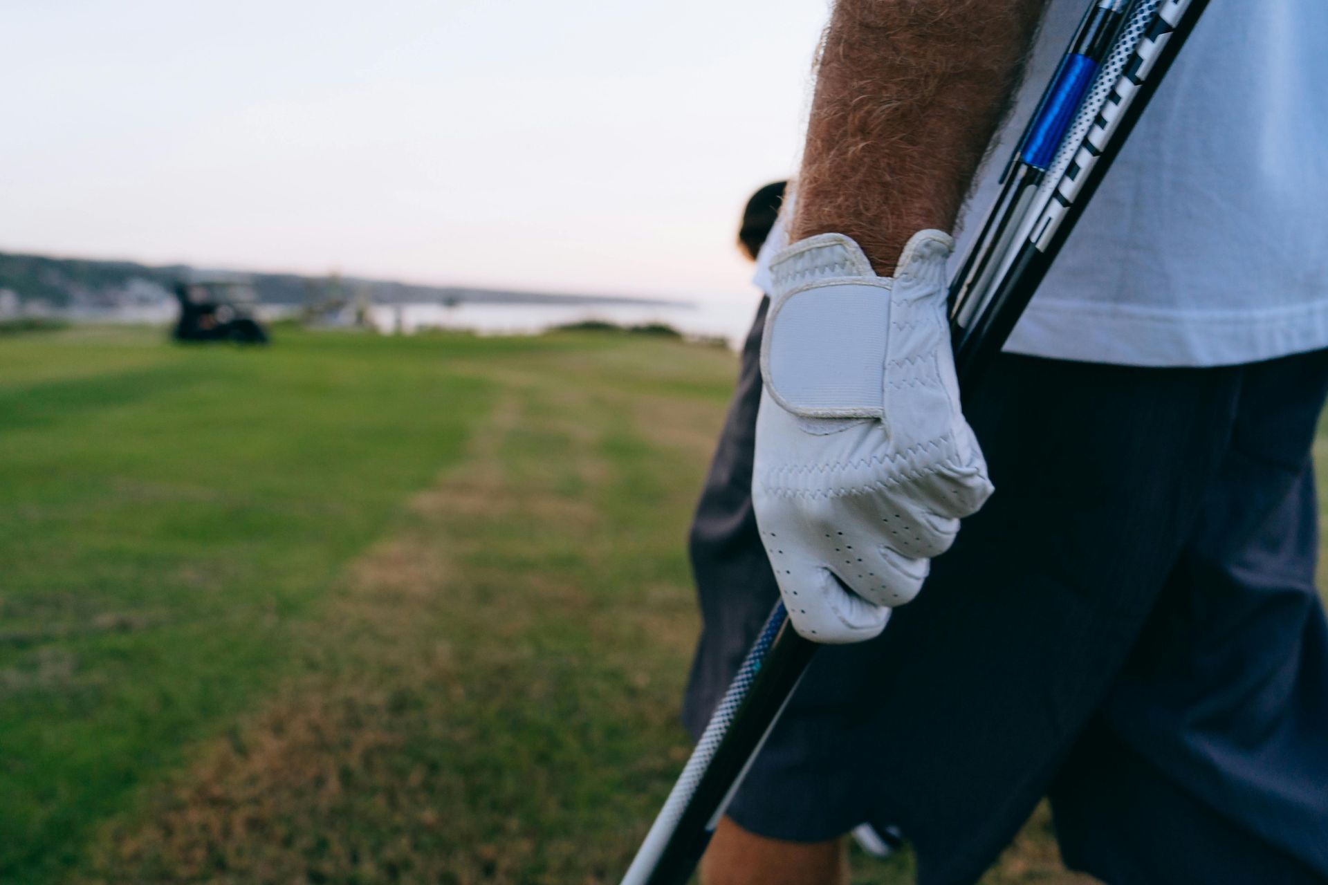 Close-up of a person wearing a white golf glove, holding a golf club on a green course at dusk.