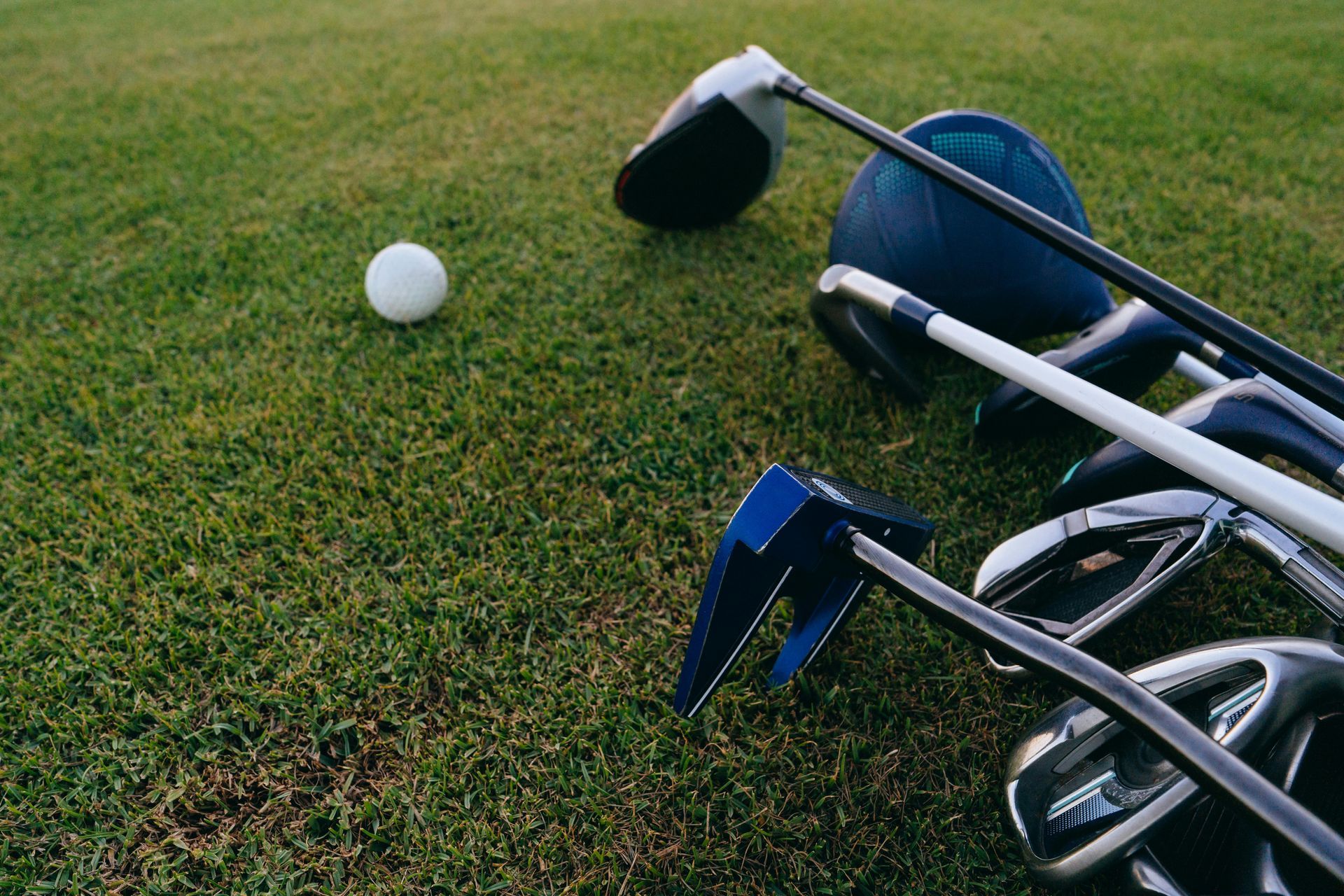 A single white golf ball rests on a green lawn beside a set of golf clubs on a bag.