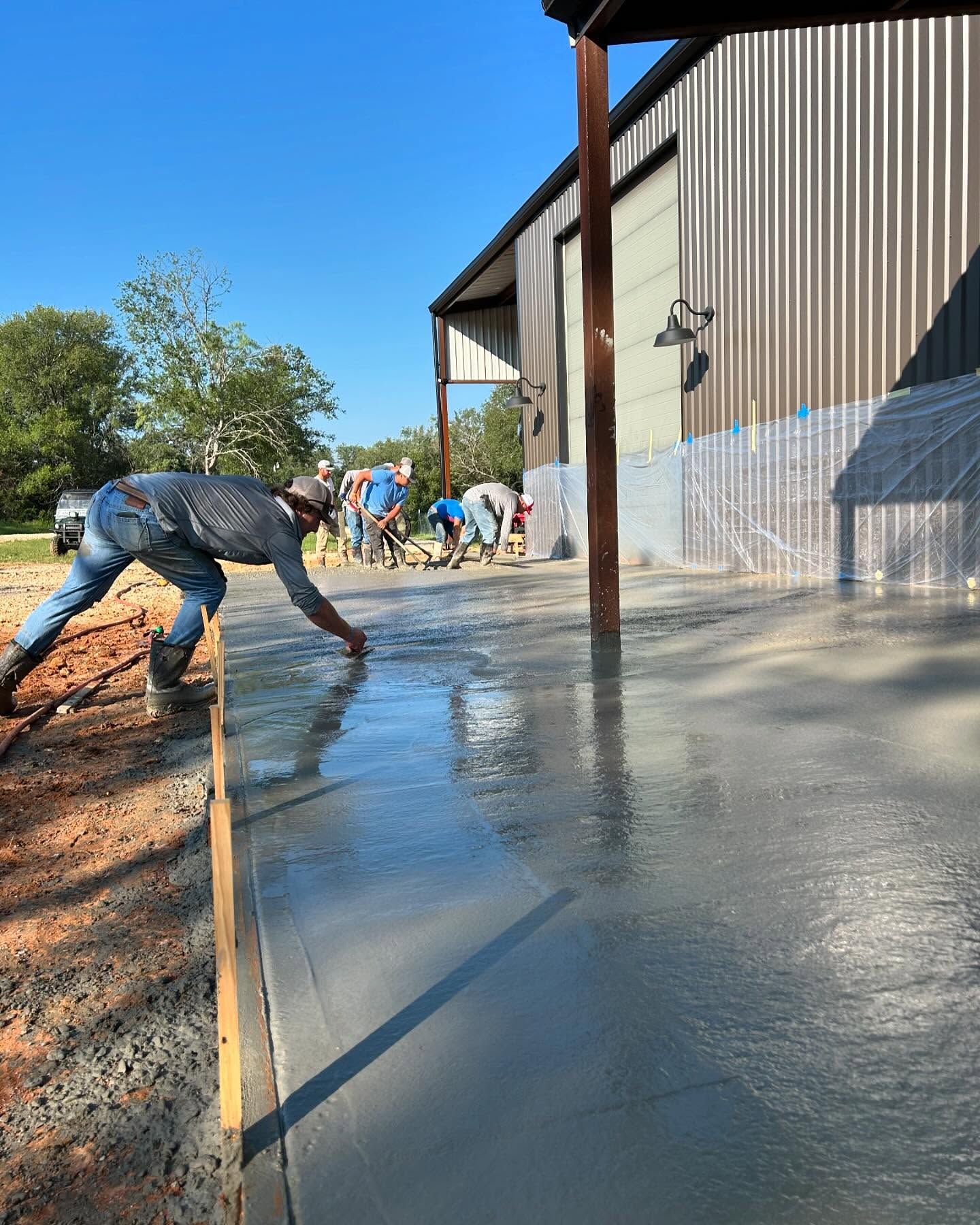 Workers pouring and smoothing concrete floor outside a metal building on a sunny day.