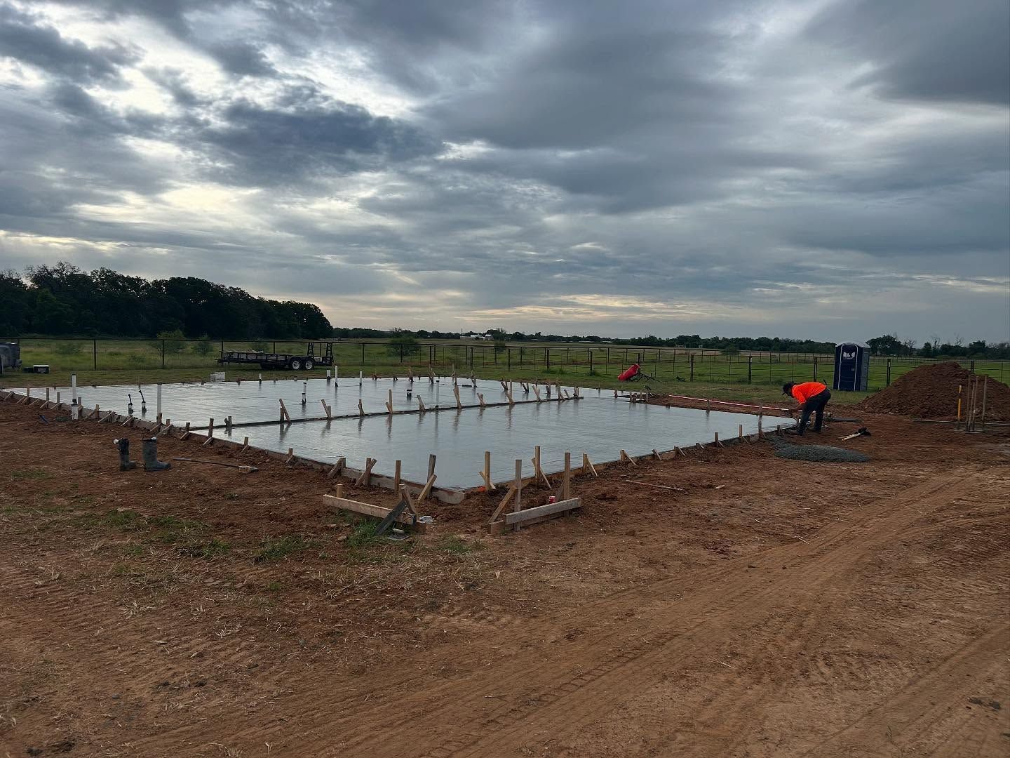Concrete foundation being poured, construction site in a field, cloudy sky.