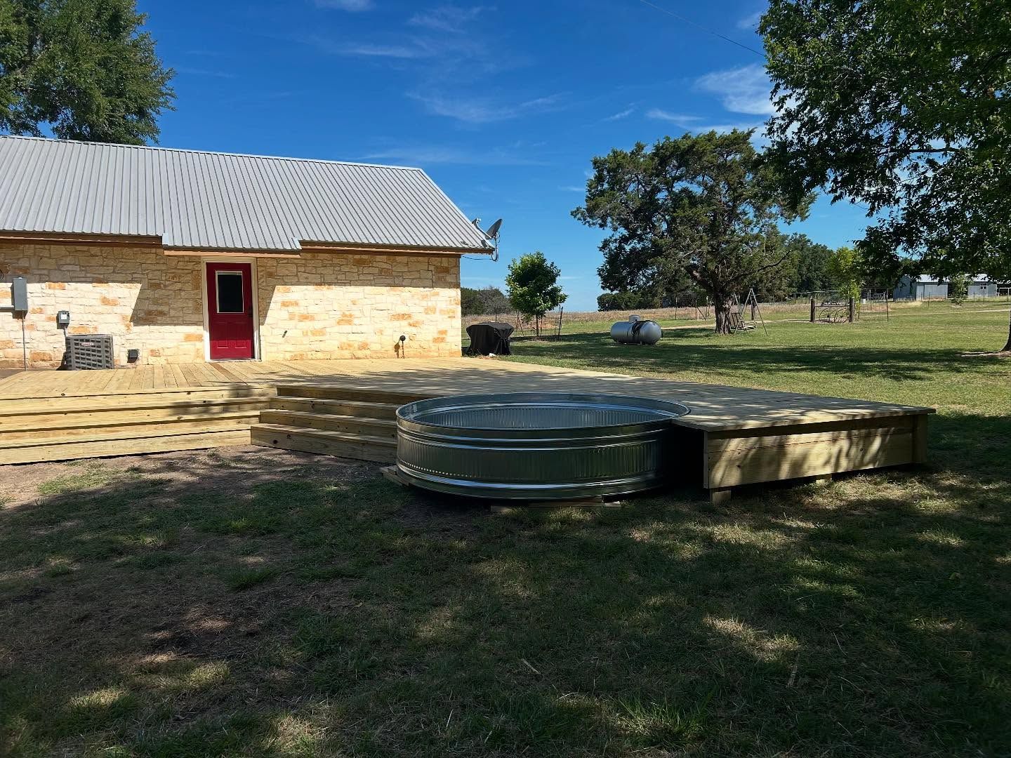 Backyard with a metal stock tank pool on a wooden deck next to a stone building with a red door.