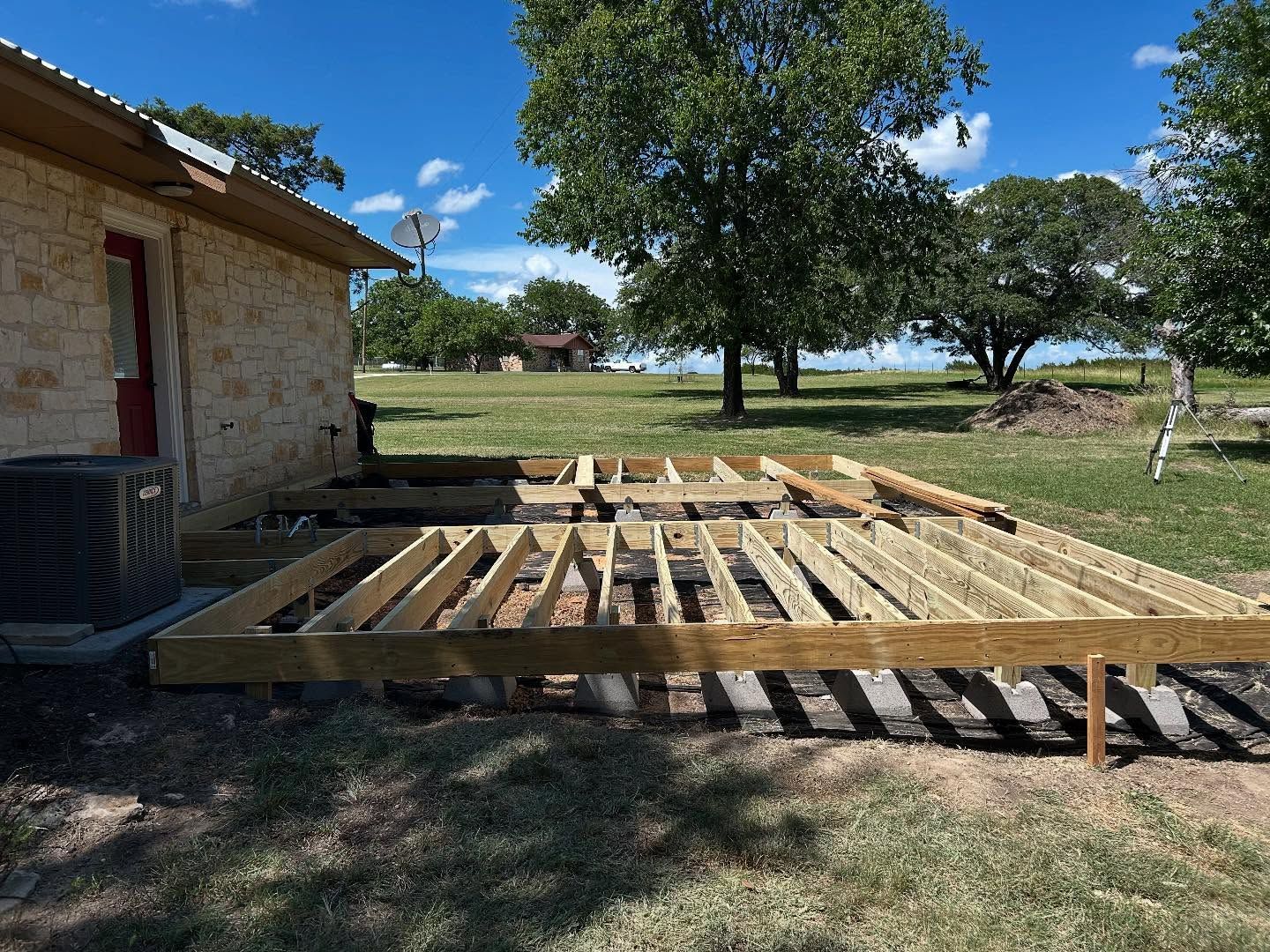 Deck construction in progress next to a tan stone building, wood framing laid out in a grassy yard.