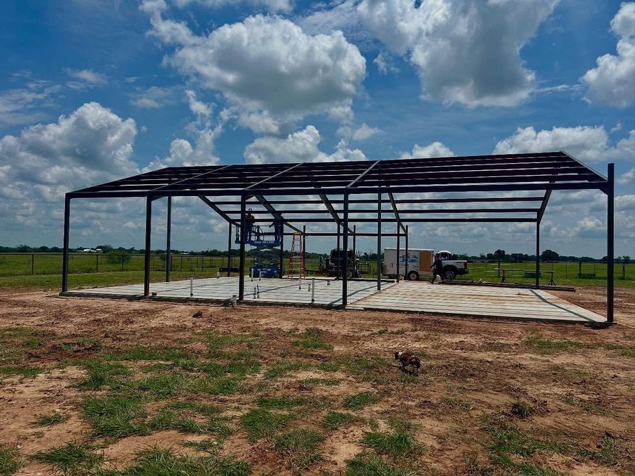 Steel frame of a large open-sided building under construction, set in a grassy field under a cloudy sky.