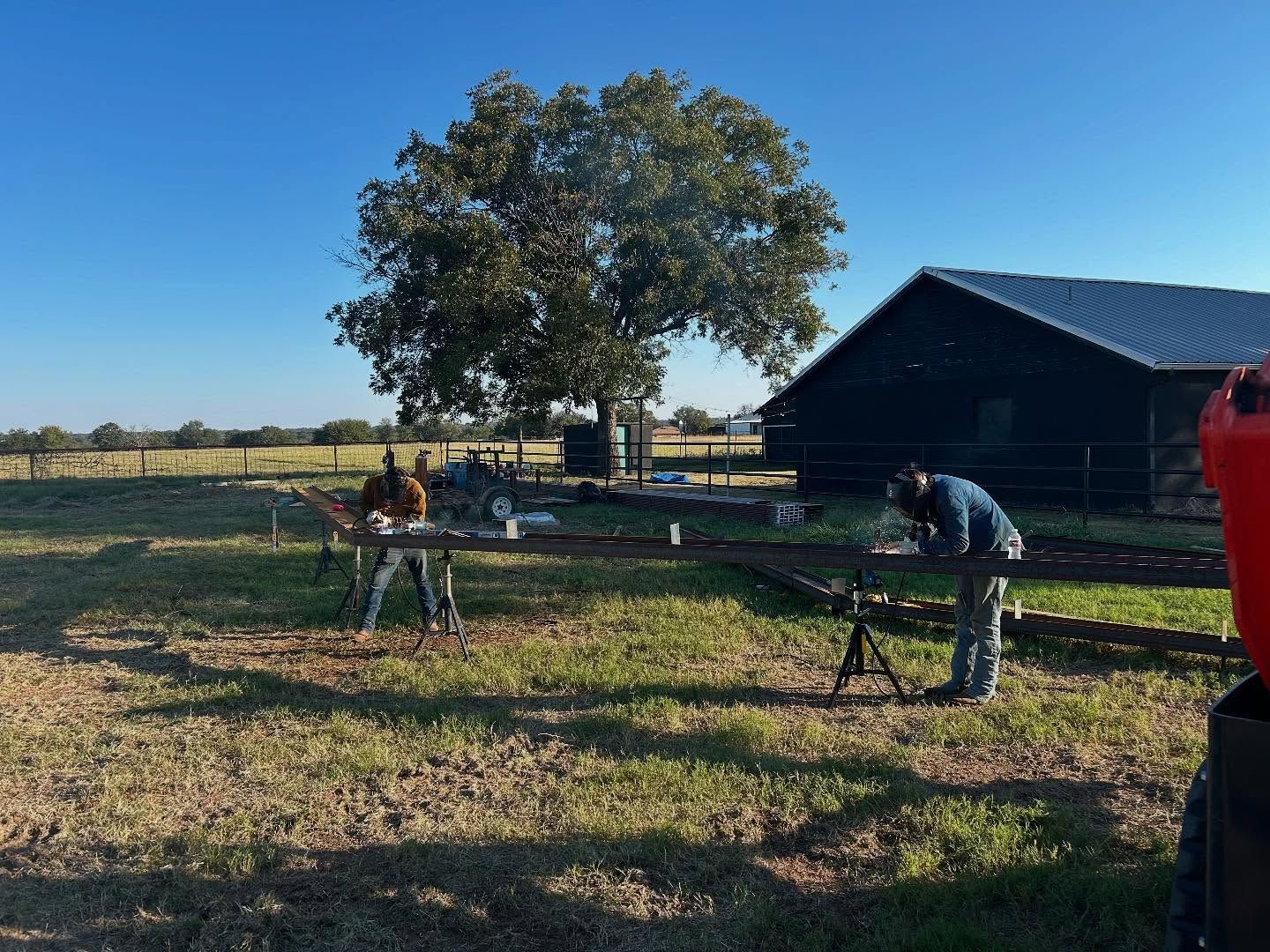 Two people working on metal outside in front of a barn and tree.