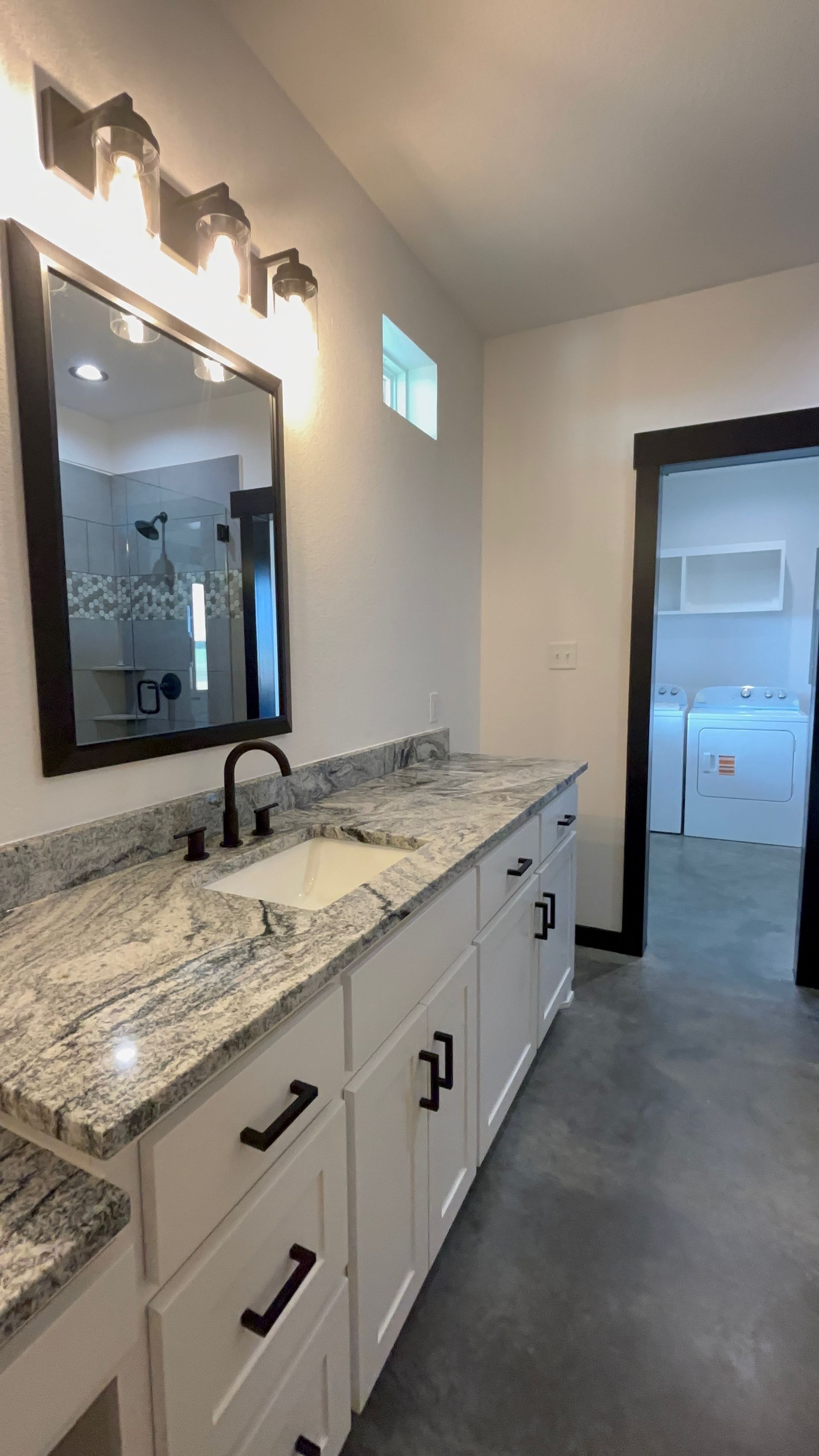 Modern bathroom with white cabinets, granite countertop, black fixtures, and a laundry room visible in the background.