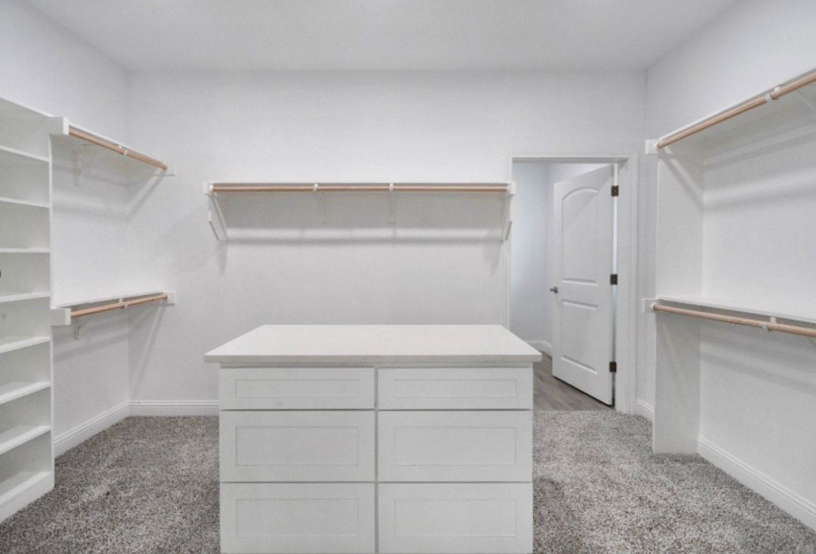 Empty white closet with central drawer unit and shelving. Doorway visible in the background.