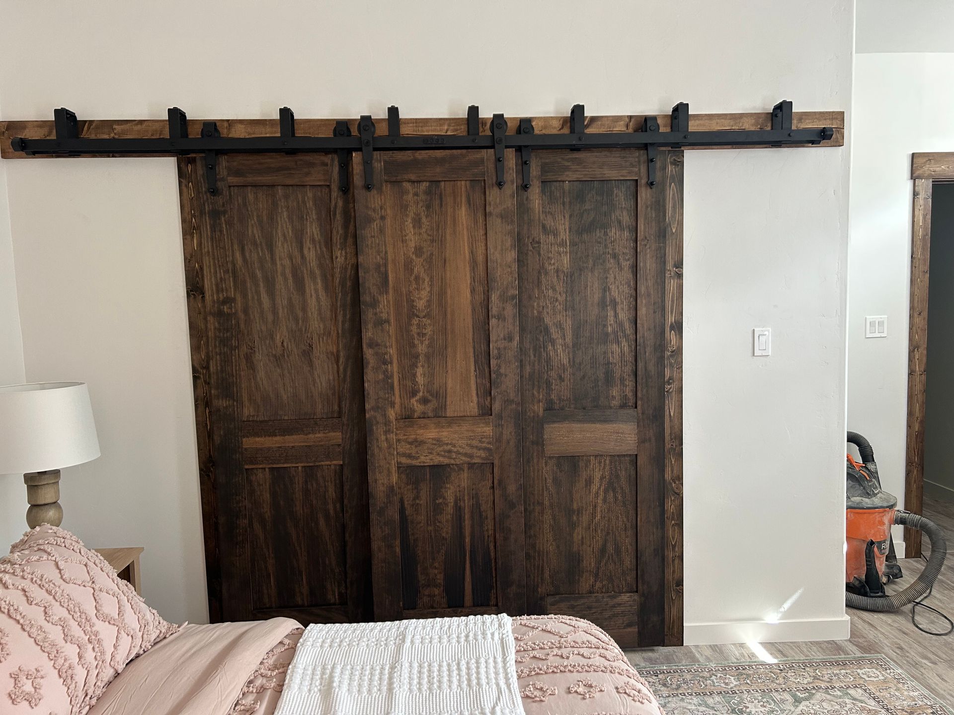 Rustic wooden barn doors on a black track above a bed. Bedroom setting, white wall, brown trim.